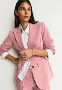 Lyserød skræddersyet blazer med hakket krave, forlommer og enkelt knaplukning, båret over en hvid skjorte.
