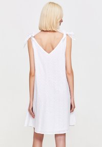 Osoba z włosami blond do ramion nosi białą, bezrękawnikową sukienkę z dekoltem w kształcie litery V i wiązanymi ramiączkami, na neutralnym tle.