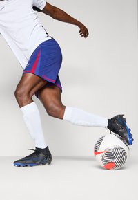 Black and blue soccer cleats, white socks, blue shorts with red accents, and a patterned soccer ball in motion on a light background.