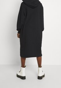 Schwarzes Kapuzen-Sweatshirt-Kleid mit langen Ärmeln, das bis zur Mitte der Wade reicht, in lässiger Passform und kombiniert mit weißen Kampfstiefeln mit schwarzen Sohlen.