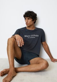 Mørk grå t-shirt med hvidt logo "Marc O'Polo" og teksten "EST. I STOCKHOLM." Bæres med matchende mørke shorts; glat stof, afslappet pasform.