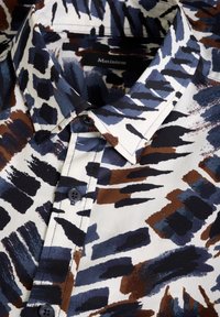 Camisa de cuello en color crema con patrones abstractos de pinceladas en azul, marrón y negro. Cuenta con botones en la parte delantera y una textura suave.