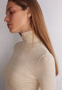 Beige rolák vyrobený z texturované tkaniny, přiléhavého střihu s vysokým límcem, s jemným, třpytivým leskem a bezšvovými rameny.