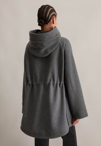 Manteau en mélange de laine gris avec une grande capuche, une taille cintrée et des manches amples. Le tissu a une texture lisse et un motif chiné subtil.