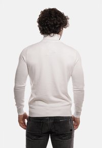 Maglione bianco a maniche lunghe con collo alto, texture a coste, design aderente e superficie liscia, indossato con jeans grigio scuro.