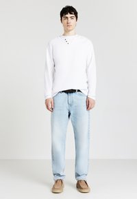 Giovane uomo in camicia bianca a maniche lunghe, jeans blu chiaro, cintura marrone e scarpe in suede color sabbia in piedi contro uno sfondo bianco uniforme.