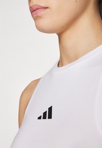 Λευκό αμάνικο μπλουζάκι με ψηλό λαιμό, που φέρει το λογότυπο adidas σε μαύρο χρώμα στο στήθος, κατασκευασμένο από μαλακό, ελαφρύ ύφασμα.