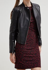 Femme portant une veste en cuir noir zippée sur une robe ajustée à motifs rouges et noirs, se tenant contre un fond uni.