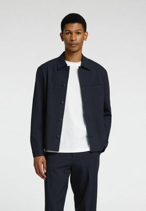 Chaqueta azul marino de botones, hecha de un tejido suave, con cuello puntiagudo y un único bolsillo en el pecho, llevada sobre una camiseta blanca.