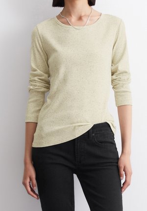 Femme portant un haut côtelé beige clair à manches longues et un jean noir, debout devant un fond blanc uni.