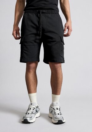 Férfi fekete oldalzsebes cargo rövidnadrágban, fehér zokniban és szürke sportcipőben, egyszínű háttér előtt áll.