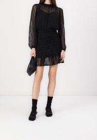 Svart klänning med genomskinliga långa ärmar, smockad midja och volangkant. Bärs med svarta strumpor och klackskor, kompletterad med en svart väska.