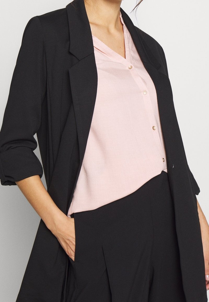 Femme portant un chemisier rose pâle boutonné, rentré dans un pantalon noir taille haute et un blazer noir avec les manches retroussées. Main dans la poche.