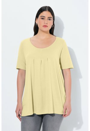 Donna con capelli lunghi e scuri che indossa una maglia a maniche corte giallo chiaro con pieghe frontali e pantaloni grigi su sfondo bianco.