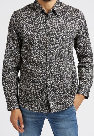 Mies, jolla on musta pitkähihainen napillinen paita, jossa on pieni valkoinen kukkakuvio, ja siniset farkut, seisoo käsi yhdessä taskussa.