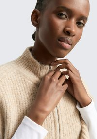 Beige gebreide zip-up trui met een hoge kraag, vervaardigd uit gestructureerde stof en een zilveren rits. Model raakt de rits met beide handen aan.