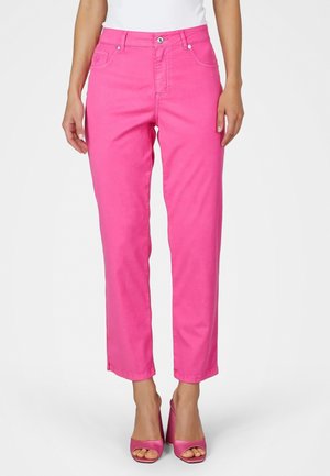 Frau trägt leuchtend pinke gerade geschnittene Hose mit vorderen Taschen und passende pinke offene High-Heel-Sandalen, steht vor einem weißen Hintergrund.