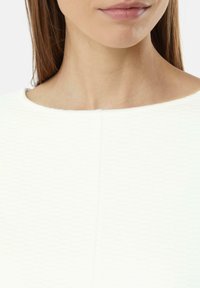 Haut blanc texturé avec un large col et de légères coutures verticales. Tissu lisse avec une apparence structurée.