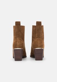 Un paio di stivali alla caviglia in suede marrone con tacco in legno blocco, mostrati da dietro su uno sfondo bianco.