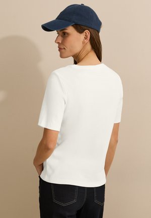 T-shirt blanc à manches courtes avec un col rond, associé à un jean foncé et une casquette de baseball bleu marine, vu d'un angle arrière.
