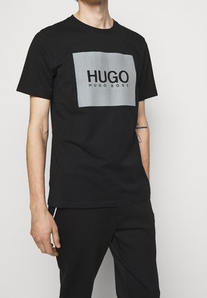 Mężczyzna noszący czarną koszulkę Hugo Boss z dużym szarym prostokątem i tekstem logo z przodu, w połączeniu z czarnymi spodniami, na jednolitym tle.