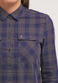 Camicia a quadri navy e marrone con colletto button-down, due tasche sul petto e bottoni blu. Presenta un tessuto leggero e testurizzato.