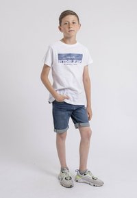 T-shirt blanc à manches courtes avec un texte graphique bleu, associé à un short en denim bleu retroussé au niveau de l'ourlet. Chaussures de sport grises avec des accents colorés.
