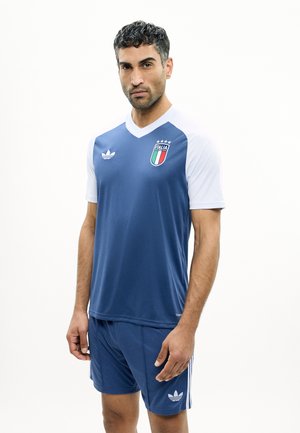 Uomo che indossa una maglia da calcio dell'Italia azzurra e bianca con pantaloncini abbinati, in piedi davanti a uno sfondo bianco semplice.