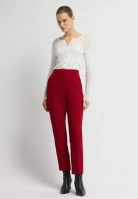 Weißes, geripptes Langarmoberteil mit Knöpfen und V-Ausschnitt, kombiniert mit hochtaillierten, roten, maßgeschneiderten Hosen und schwarzen Stiefeletten.