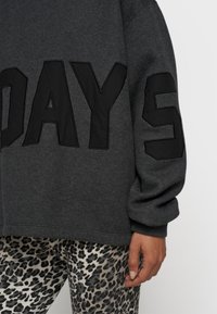 Grauer übergroßer Pullover mit großen schwarzen Buchstaben, die "DAYS" schreiben, kombiniert mit beigen Leggings im Leopardenmuster. Weicher Stoff, entspannter Schnitt.