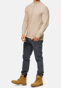 Hombre vestido con un suéter beige con cremallera, jeans oscuros de corte ajustado y botas de trabajo de cuero color marrón claro, de pie con una mano en el bolsillo.