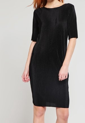 Kobieta ubrana w czarną, teksturowaną sukienkę o długości do kolan i krótkich rękawach, stojąca na tle jednolitego jasnoszarego tła.