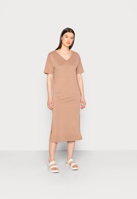 Vestido de mangas curtas em V, na cor castanho claro, feito de um tecido suave com fendas laterais. Combinado com sandálias de plataforma brancas.