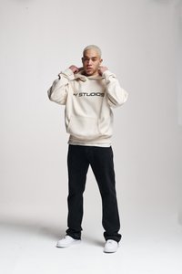Hanorac crem cu inscripția "STUDIOS" imprimată în negru, buzunar frontal, asociat cu pantaloni negri și adidași albi. Design simplu și casual.