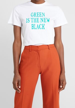T-shirt blanc avec texte teal « GREEN IS THE NEW BLACK » porté avec un pantalon orange taille haute, mains détendues le long du corps.