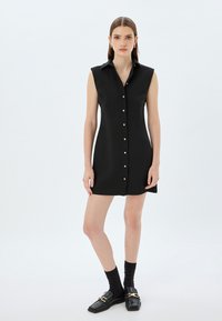 Zwart mouwloos jurk met een knoopsluiting aan de voorkant, puntige kraag en gouden hardware-accenten. Gladde textuur en aansluitende silhoutte.