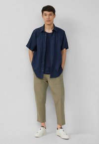 Navy korte mouwen shirt, knoopjes, gedragen over een donkerblauwe top; gecombineerd met lichtolijfgroene tapered broek en witte sneakers. Minimalistisch ontwerp.