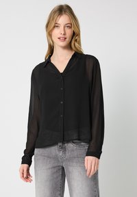 Blusa negra transparente con botones en el frontal y cuello. Cuenta con mangas largas y un diseño ligeramente corto. Combinada con jeans grises.