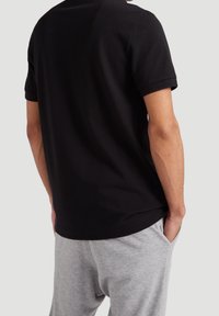 Muž stojí s rukama v kapsách, oblečený v černém polo tričku s krátkým rukávem a světle šedých teplácích, na jednobarevném pozadí.