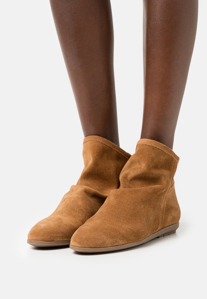 Brązowe zamszowe botki za kostkę o miękkiej teksturze, z projektowanym złożeniem, spiczastym noskiem i minimalistyczną gumową podeszwą, umieszczone w pobliżu nagich nóg.