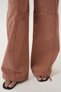 Wide-leg pants in a rust color made from textured fabric, featuring visible stitching and a relaxed fit, paired with black sandals.