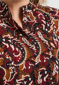 Un tessuto marrone con motivo paisley presenta un colletto arricciato e bottoni, con colori che includono rosso scuro, bianco e accenti verdi.