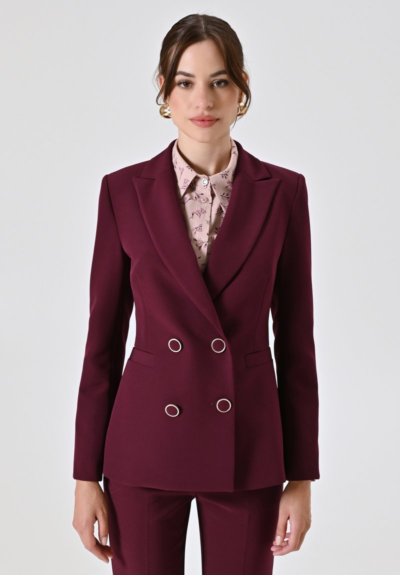 Blazer doppiopetto bordeaux con accessori in argento, rever a punta e due tasche, indossato su una blouse chiara a fantasia. Tessuto dalla consistenza liscia.
