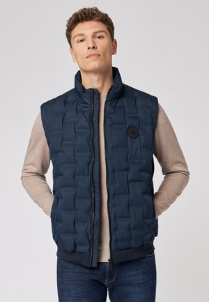 Navy blue, sleeveless puffer vest with a zip front, padded texture, and a round black logo on the chest. Worn over a light sweater.