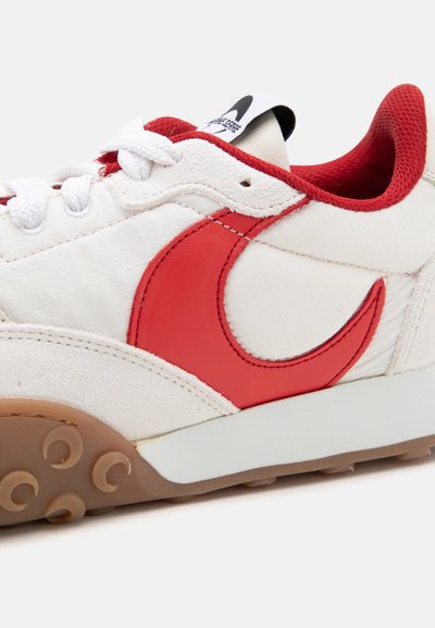 White sneaker featuring a red swoosh, textured upper, padded collar, and a gum rubber sole with circular patterns.
