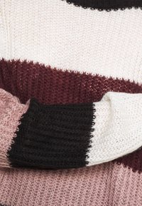 Pletený svetr s horizontálními pruhy v odstínech černé, bílé, bordó a růžové; různorodé textury a volný pletený design.