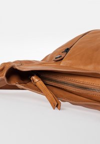 Bruine leren tas met een zacht gebogen vorm, een ritsvak aan de binnenkant en een verstelbare schouderriem. Zichtbare stiksels en ritsdetails.