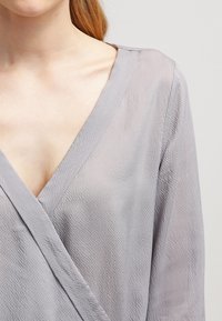 Top avvolgente grigio chiaro, con texture, scollatura a V profonda e maniche lunghe, caratterizzato da una leggera lucentezza e dettagli di design minimalisti.