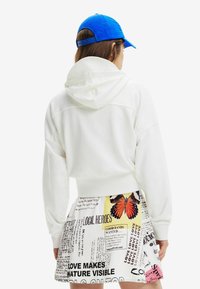 Cropped hoodie bianco con cappuccio, abbinato a una gonna stampata multicolore con testi e motivi di farfalle. Un cappellino blu completa l'outfit.
