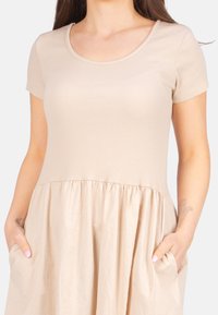 Beiges Kleid mit einem gerippten Oberteil, kurzen Ärmeln und einem gerafften, fließenden Rock. Das Design umfasst zwei seitliche Taschen.